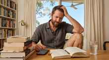 Stressed man at desk tracking his reading stats on his phone and in a notebook